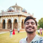 Young man takes a cheerful selfie at Humayun's Tomb, showcasing historic Mughal architecture.