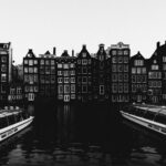 Black and white view of Amsterdam's canal houses with two boats and reflections.